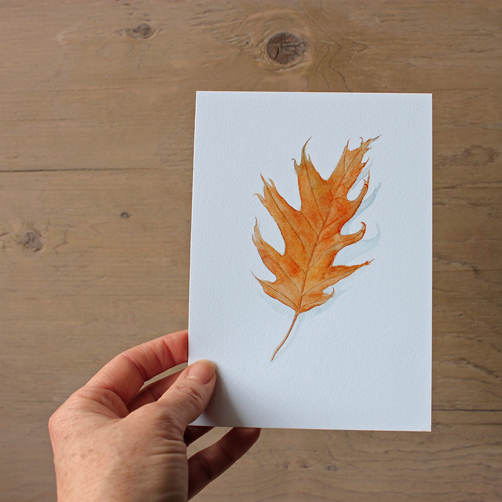 An autumn art print featuring a watercolor painting of an orange oak leaf. Hand of artist Kathleen Maunder.