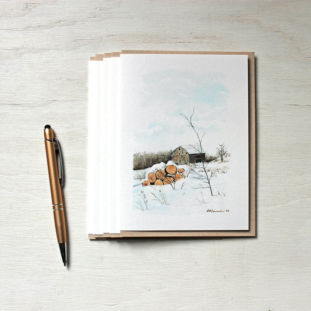 A set of five note cards featuring a watercolor painting of a winter rural scene with a rustic barn and a pile of firelogs covered with snow. Artist Kathleen Maunder.