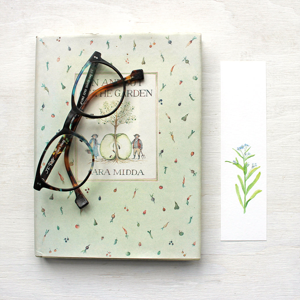 A paper bookmark featuring a watercolor painting of a stem of blue forget me nots. Artist Kathleen Maunder.