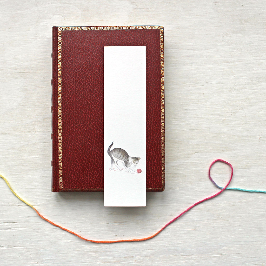 A paper bookmark featuring a watercolor painting of a tabby kitten playing with a ball of yarn. Artist Kathleen Maunder.