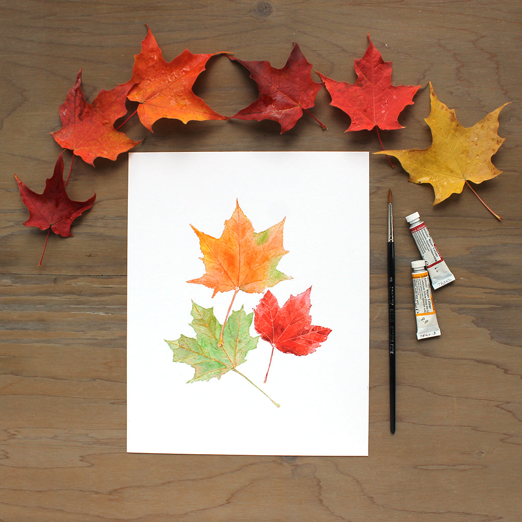An art print featuring a detailed watercolor painting of three maples leaves. Orange, red and green tones. Artist Kathleen Maunder.