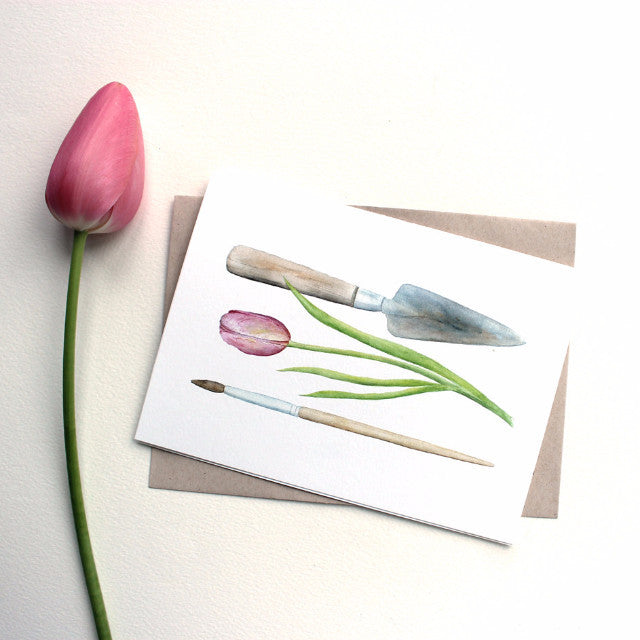 A note card featuring a watercolor painting of a garden trowel, a pink tulip and an artist's paintbrush. Artist Kathleen Maunder.