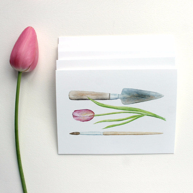 A note card featuring a watercolor painting of a garden trowel, a pink tulip and an artist's paintbrush. Artist Kathleen Maunder.