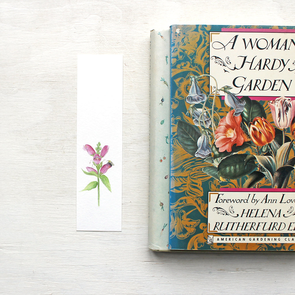 A floral bookmark featuring a watercolor painting of pink turtlehead and a bee. Artist Kathleen Maunder.