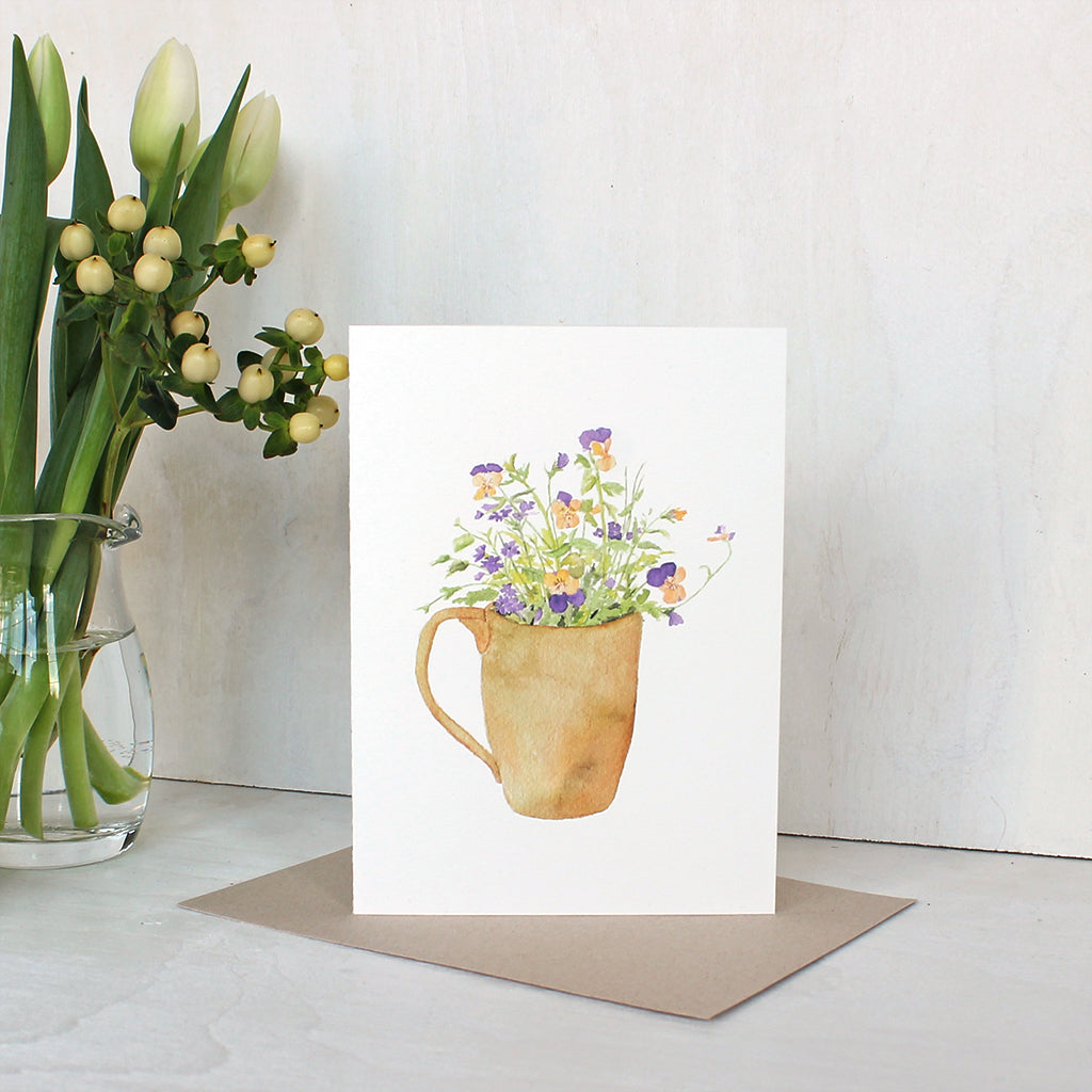 A note card featuring a watercolor painting of a mug filled with yellow and purple violas as well as purple verbena. Artist Kathleen Maunder