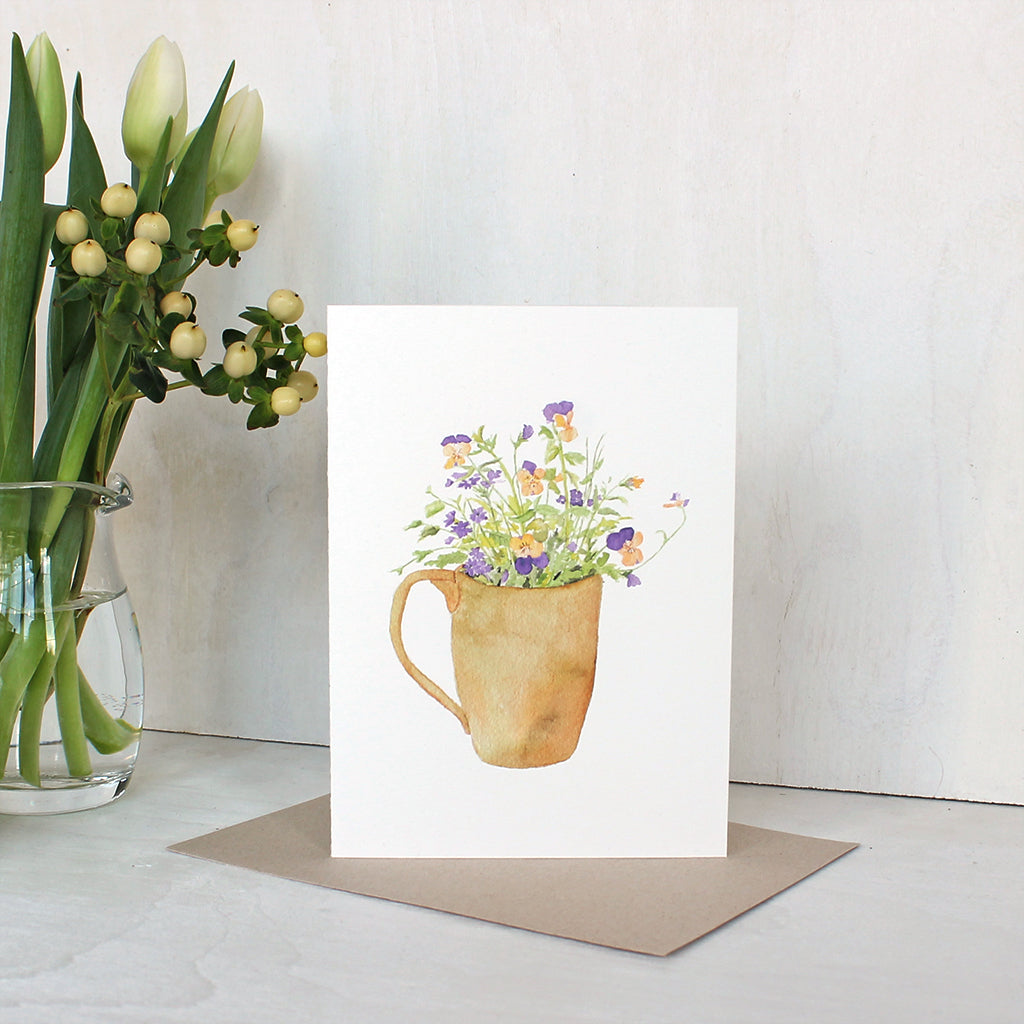 A note card featuring a watercolor painting of a mug filled with yellow and purple violas as well as purple verbena. Artist Kathleen Maunder