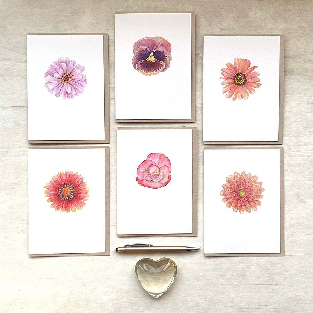 A botanical card assortment including six cards featuring single blossom paintings by Canadian watercolour artist Kathleen Maunder. At the base of the photo is a gold pen and a glass heart.