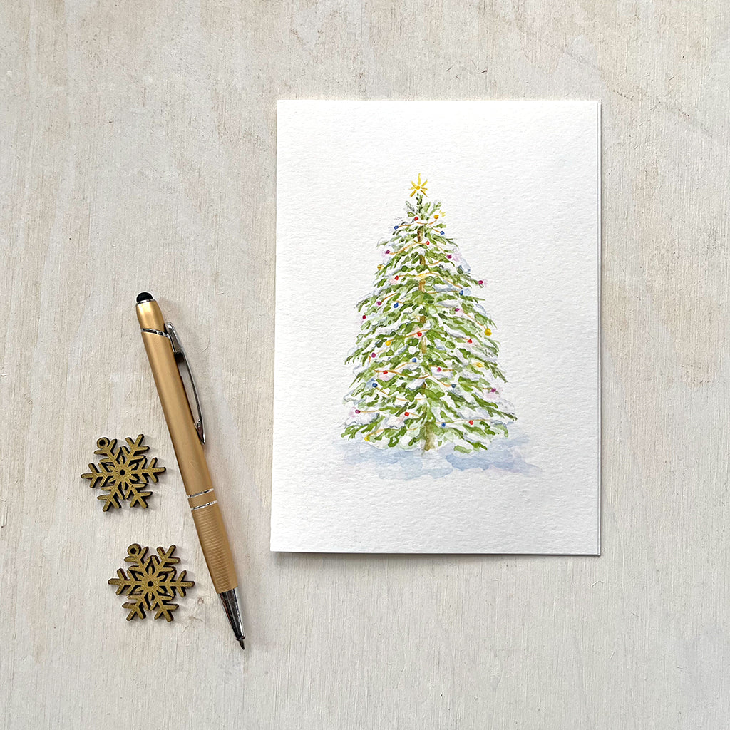 A lovely holiday card featuring a watercolor painting of a snow-covered Christmas tree adorned with colorful, sparkling lights. Artist Kathleen Maunder.