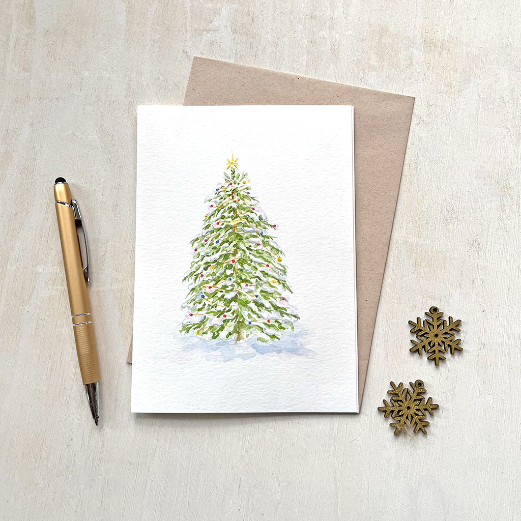 A cheerful holiday card featuring a watercolor painting of a snow-covered evergreen tree adorned with twinkly lights . Artist Kathleen Maunder.