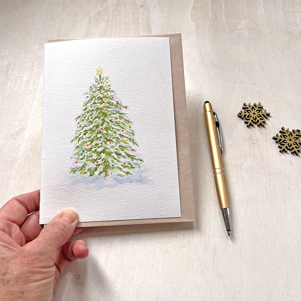 A watercolor holiday card featuring a delicate painting of a snow-covered Christmas tree adorned with colorful, sparkling lights. Artist Kathleen Maunder.