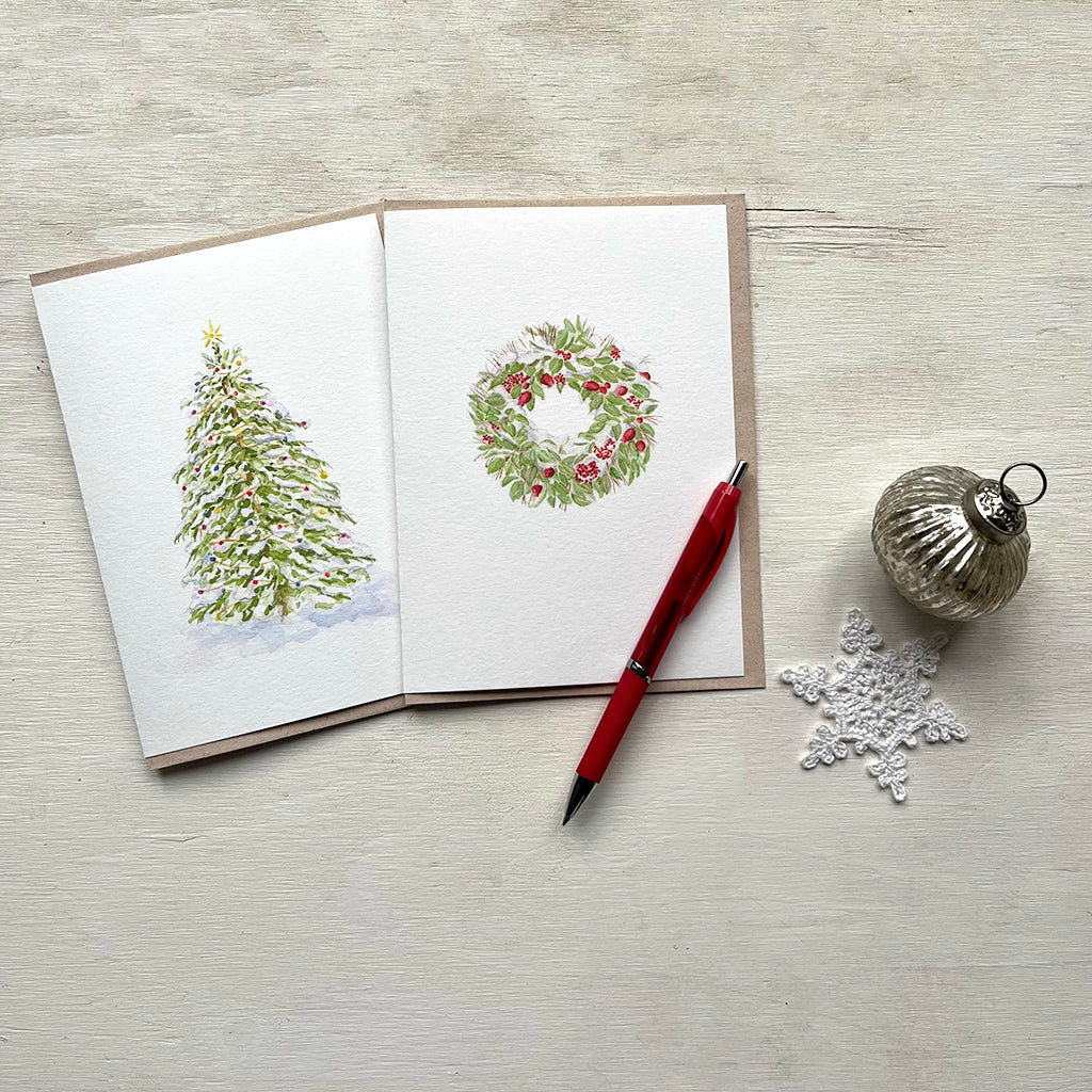 Two holiday cards featuring watercolour paintings, one a Christmas tree covered in lights and the other a snow-covered wreath. There is also a red pen, crocheted snowflake and silver ornament in the photo.