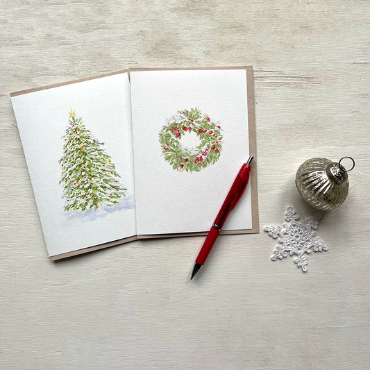 Two holiday cards featuring watercolour paintings, one a Christmas tree covered in lights and the other a snow-covered wreath. There is also a red pen, crocheted snowflake and silver ornament in the photo.