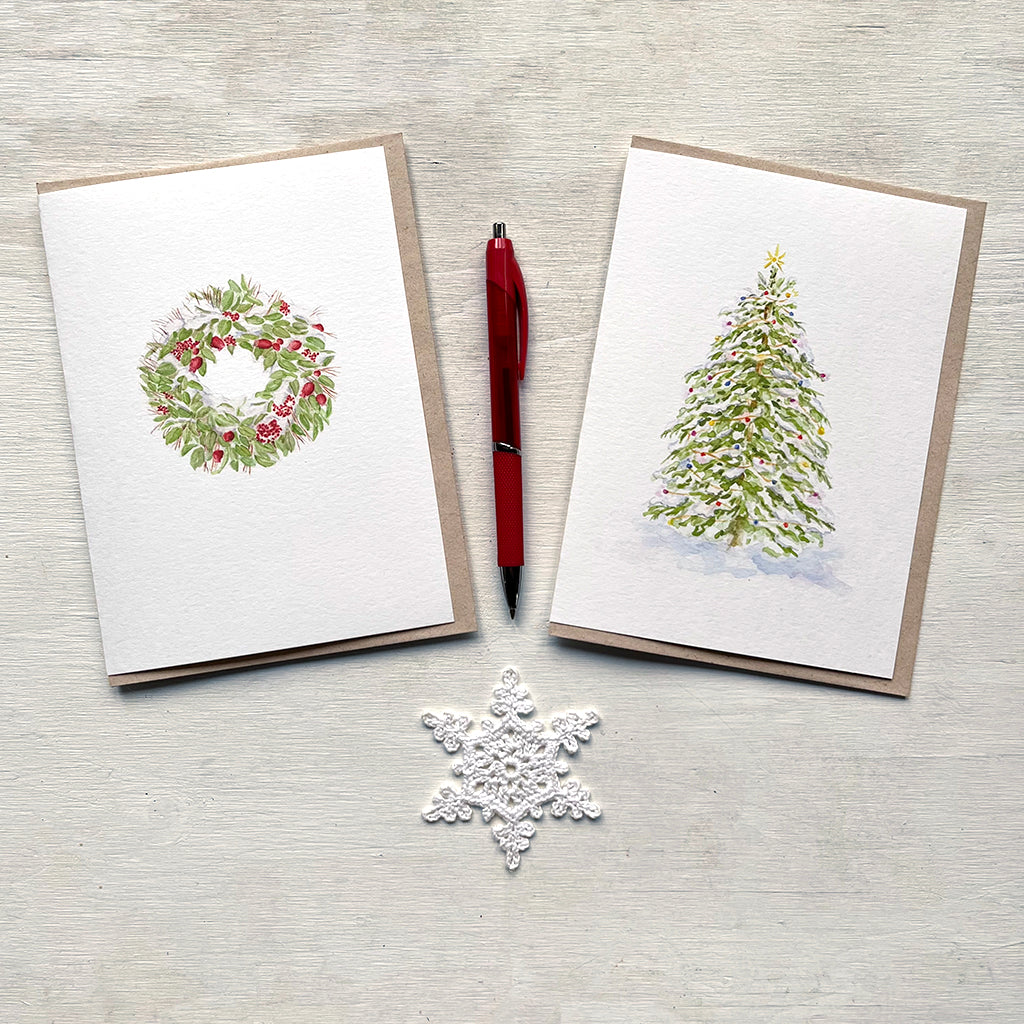 Two holiday cards featuring watercolour paintings by Canadian artist Kathleen Maunder. On the left a snow-covered wreath and on the right a Christmas tree covered in lights. There is also a red pen, and crocheted snowflake in the centre of the photo.