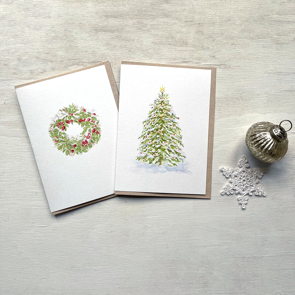 Two holiday cards featuring watercolour paintings by Canadian artist Kathleen Maunder. On the left a snow-covered wreath and on the right a Christmas tree covered in lights. There is a crocheted snowflake and a silver ornament at the right of the photo.