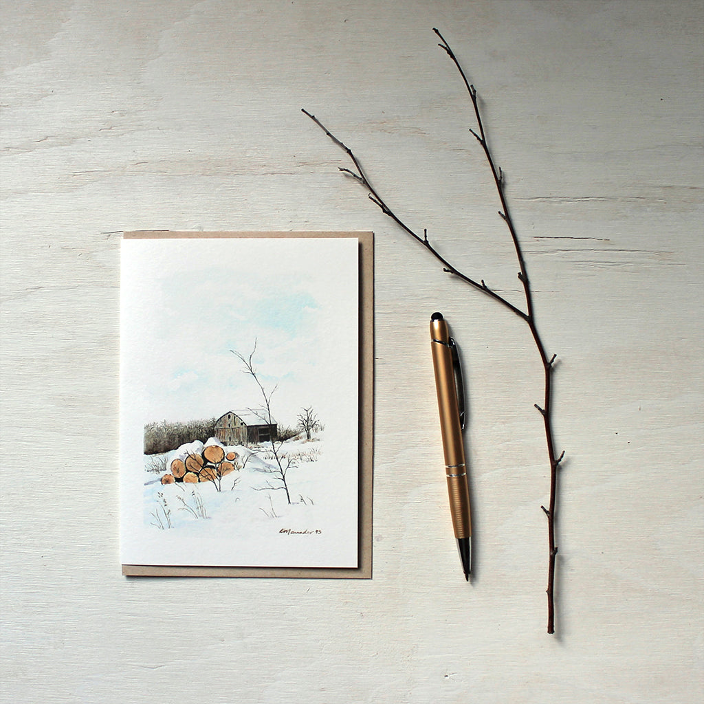 A note card featuring a watercolor painting of a winter rural scene with a rustic barn and a pile of firelogs covered with snow. Artist Kathleen Maunder.