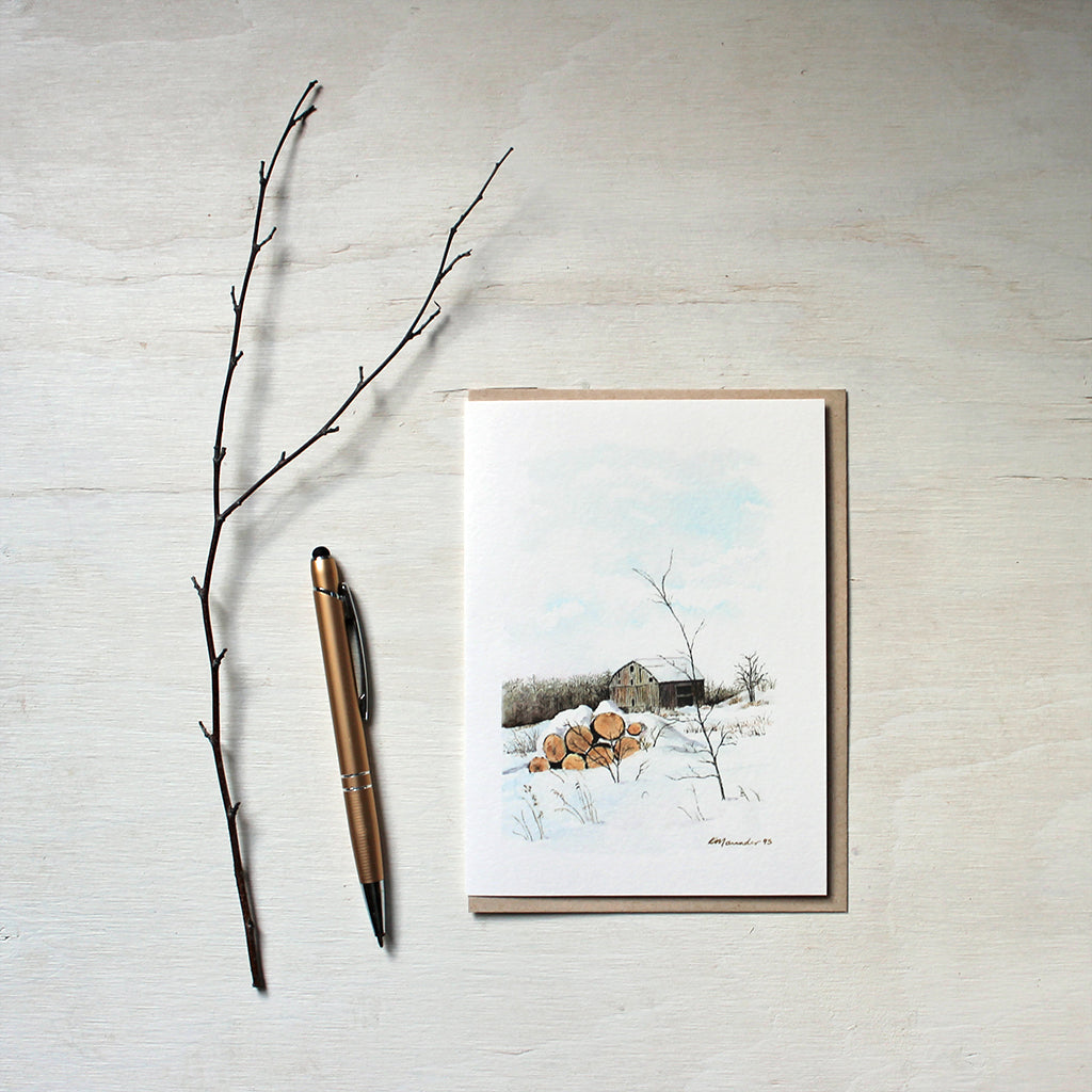 A note card featuring a watercolour painting of a winter rural scene with a rustic barn and a pile of logs covered with snow. Artist Kathleen Maunder.