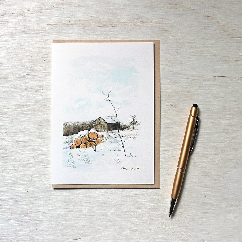 A note card featuring a watercolor painting of a winter rural scene with a rustic barn and a pile of firelogs covered with snow. Artist Kathleen Maunder.
