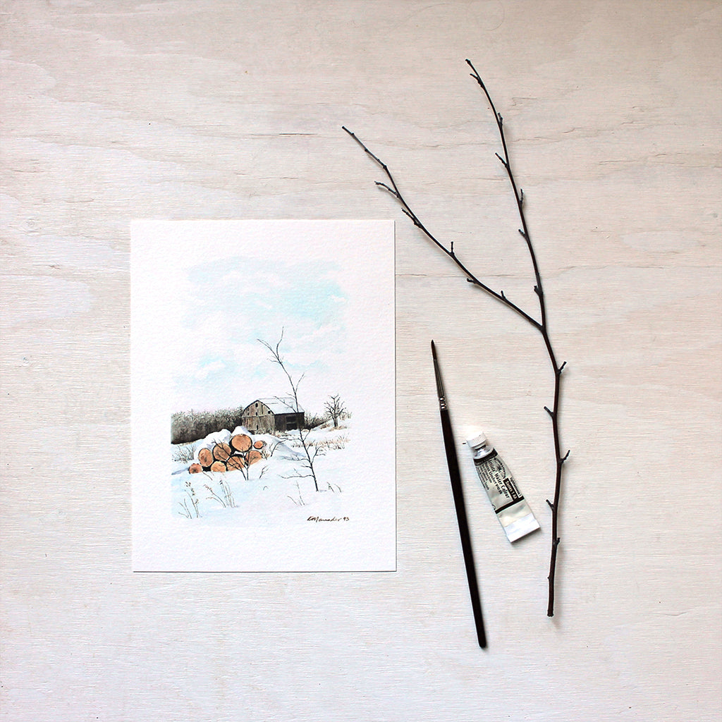 An art print of a watercolor painting of a rural winter scene including a barn and stack of logs. Artist Kathleen Maunder.