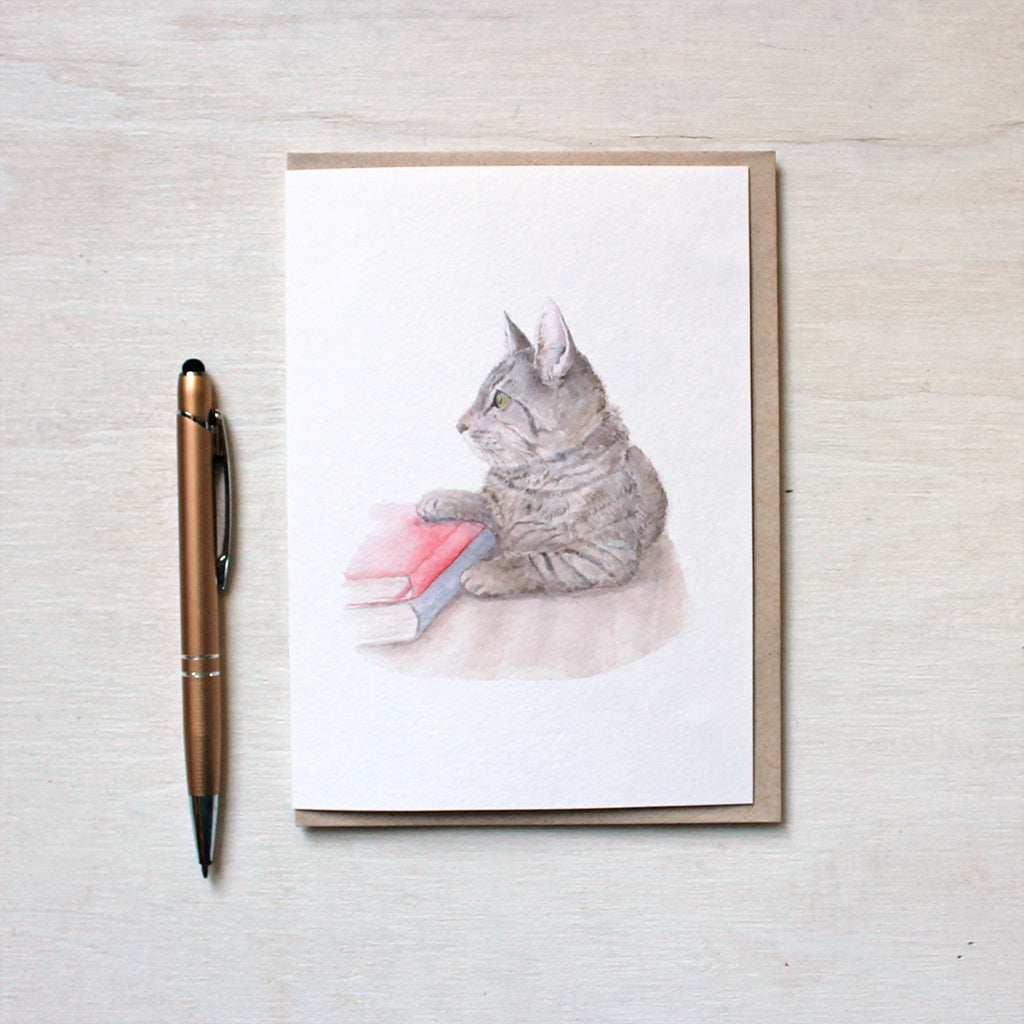 Note card featuring a watercolor painting of a tabby kitten laying beside two books. Artist Kathleen Maunder.