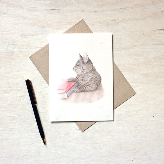 Note card featuring a watercolor painting of a sweet tabby kitten laying beside two books. Artist Kathleen Maunder.