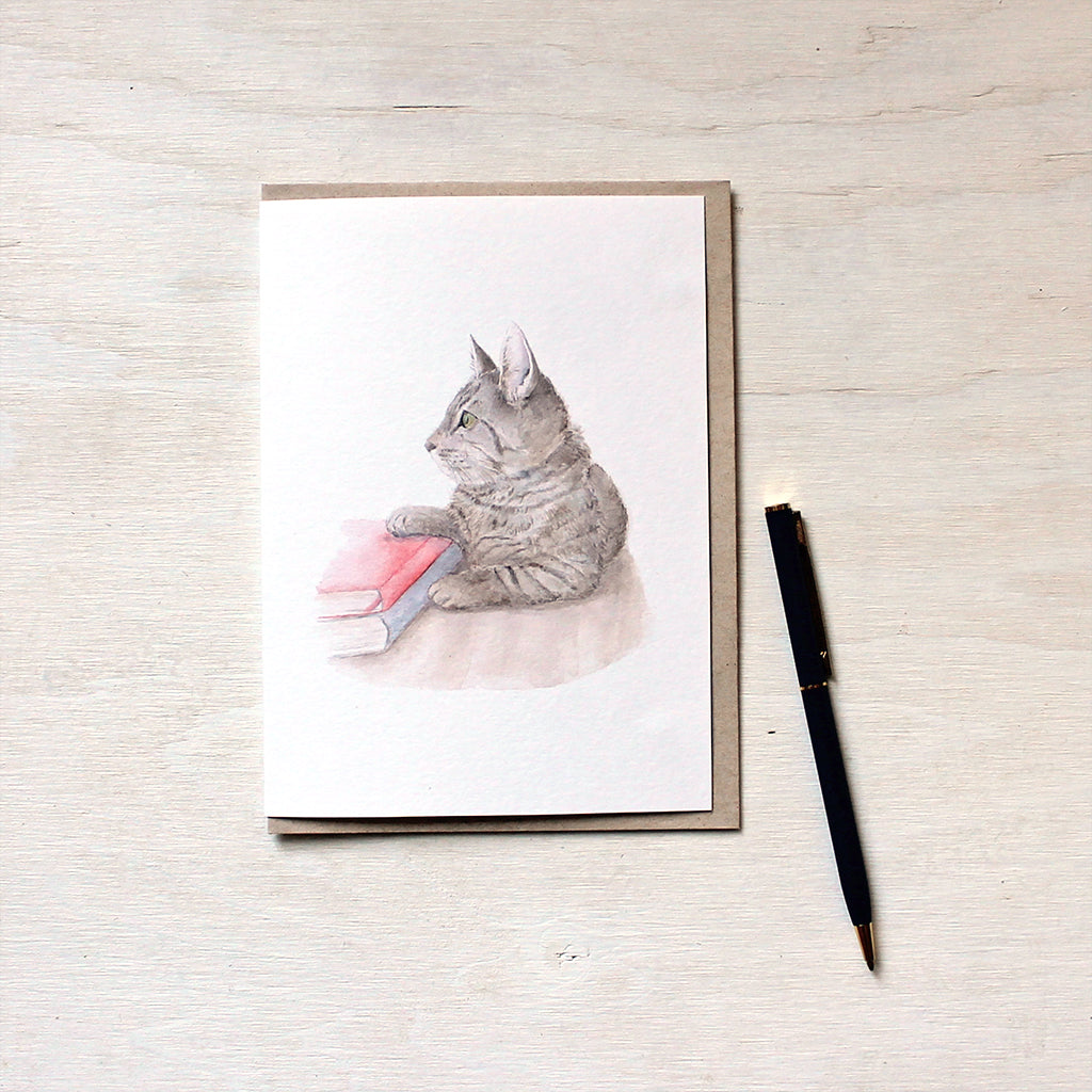 Note card featuring a watercolour painting of a sweet tabby kitten laying beside a red and blue book. Artist Kathleen Maunder.