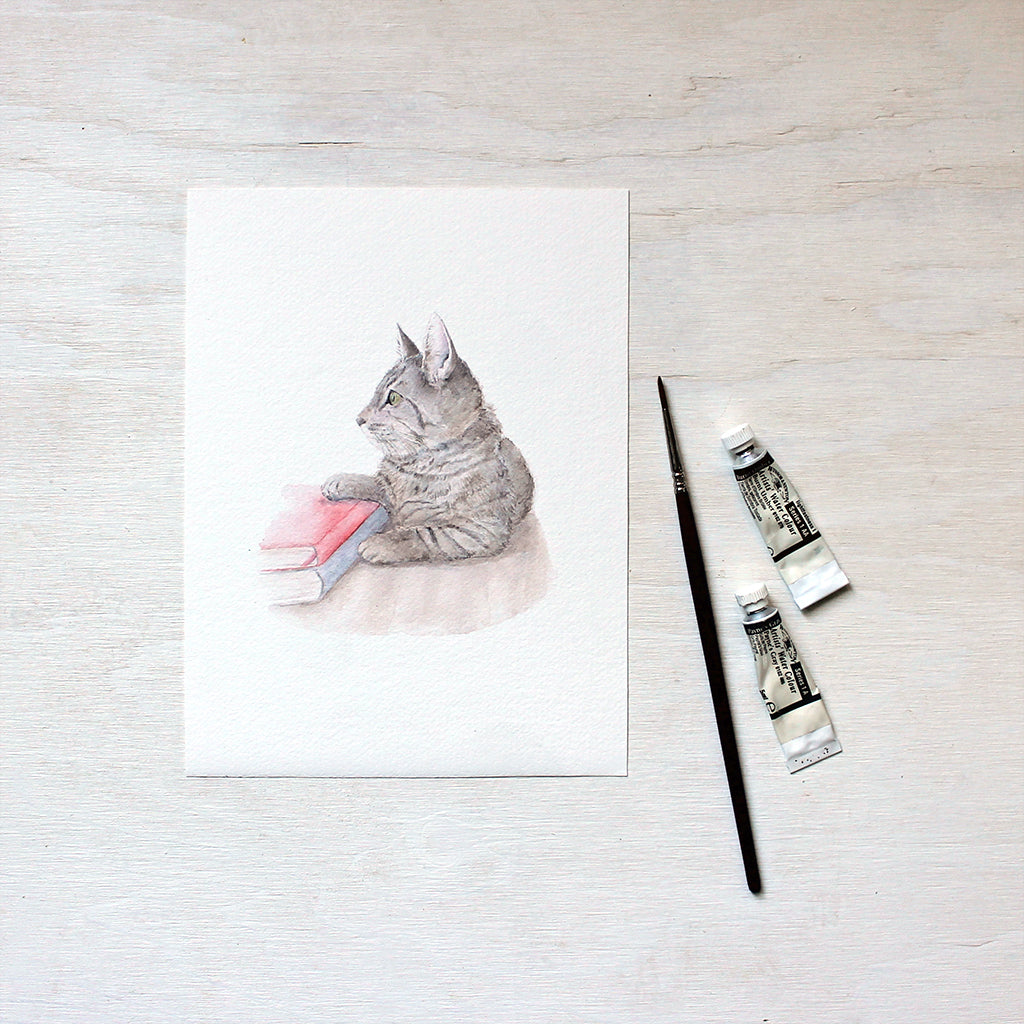 An art print featuring a watercolor painting of a sweet tabby kitten laying beside two books. Artist Kathleen Maunder.