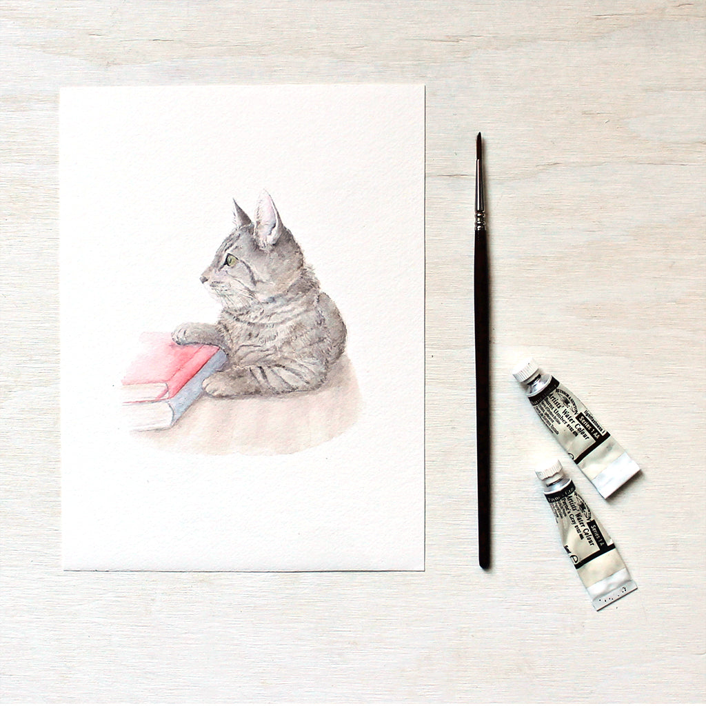 An art print featuring a watercolor painting of a sweet tabby kitten laying beside two books. Artist Kathleen Maunder.