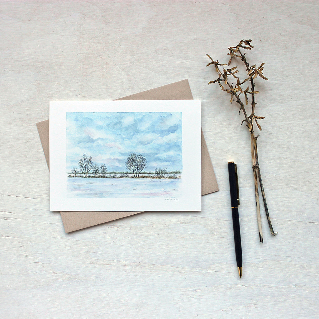 Note card featuring a watercolor painting of a cloudy sky, snowy field, a line of trees and a tiny bird. Artist Kathleen Maunder.