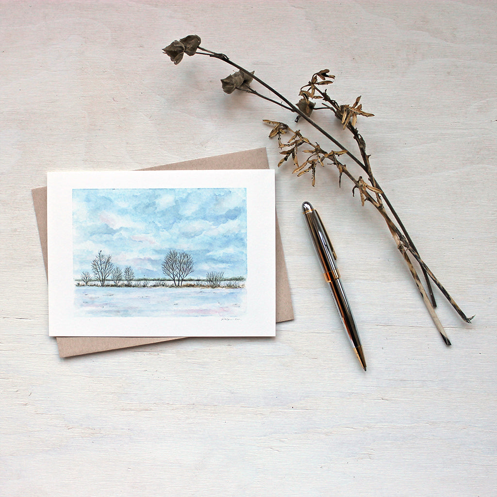 Note card featuring a watercolour painting of a cloudy sky, snowy field, a line of trees and a tiny bird. Artist Kathleen Maunder.
