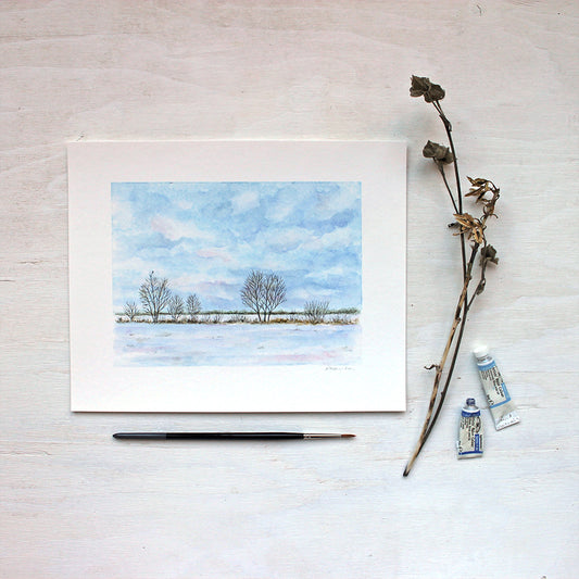 A watercolor art print featuring a wintry landscape including a rural field, trees and a tiny bird. Artist Kathleen Maunder.