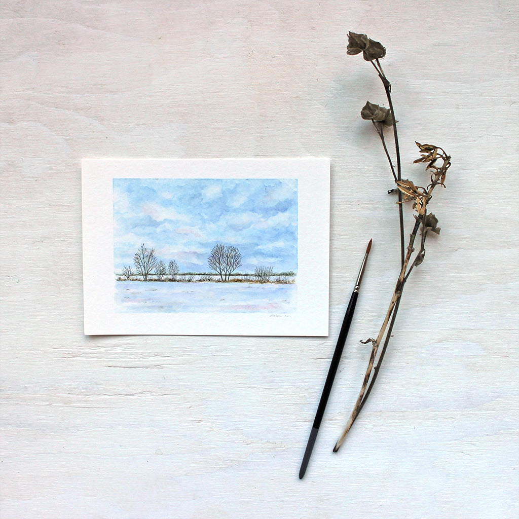 Watercolor art print featuring a painting of a snow-covered field, a tree line and a tiny bird. Artist Kathleen Maunder.