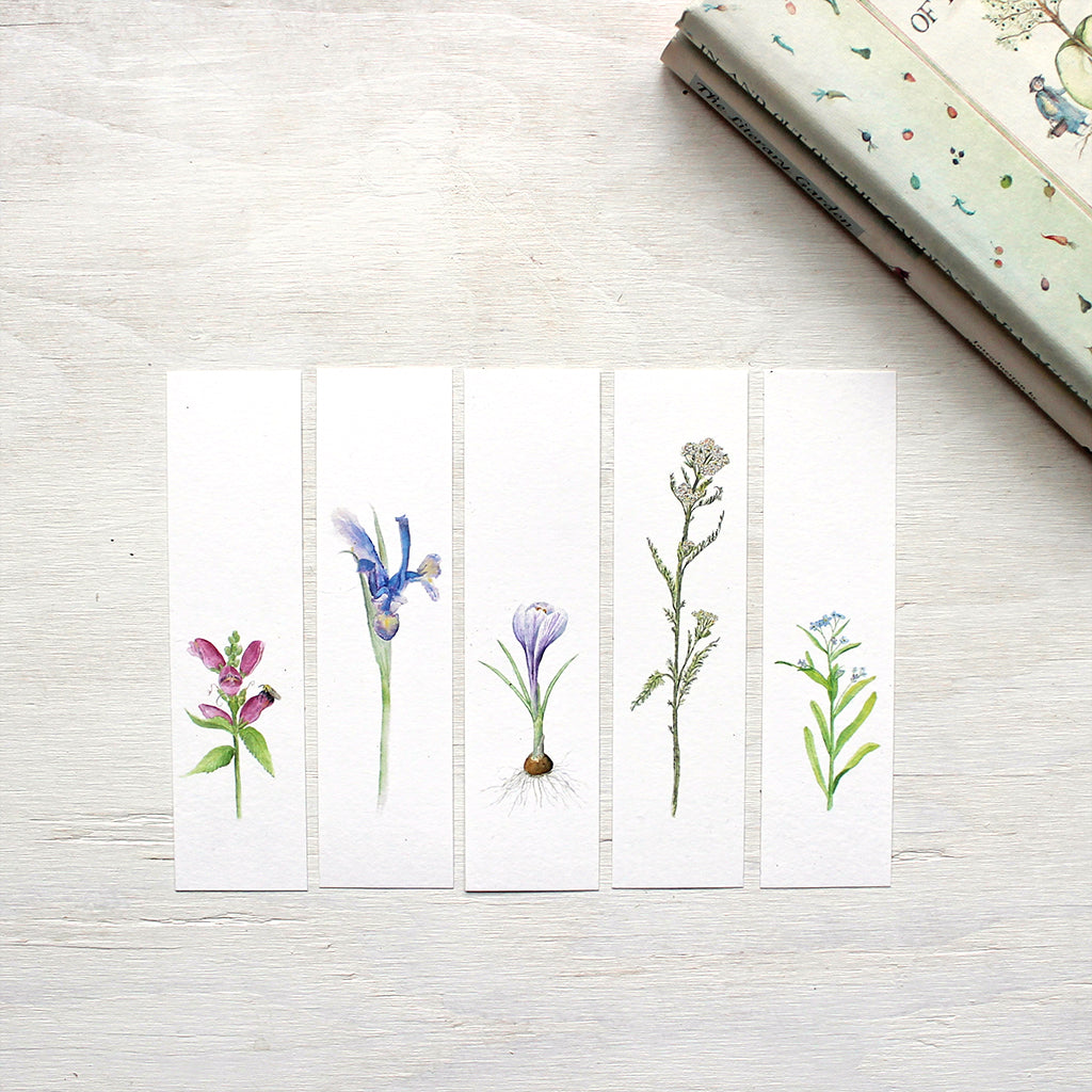 Five floral bookmarks featuring watercolour paintings of turtlehead, an iris, a purple crocus bulb, yarrow and forget me nots. Artist Kathleen Maunder