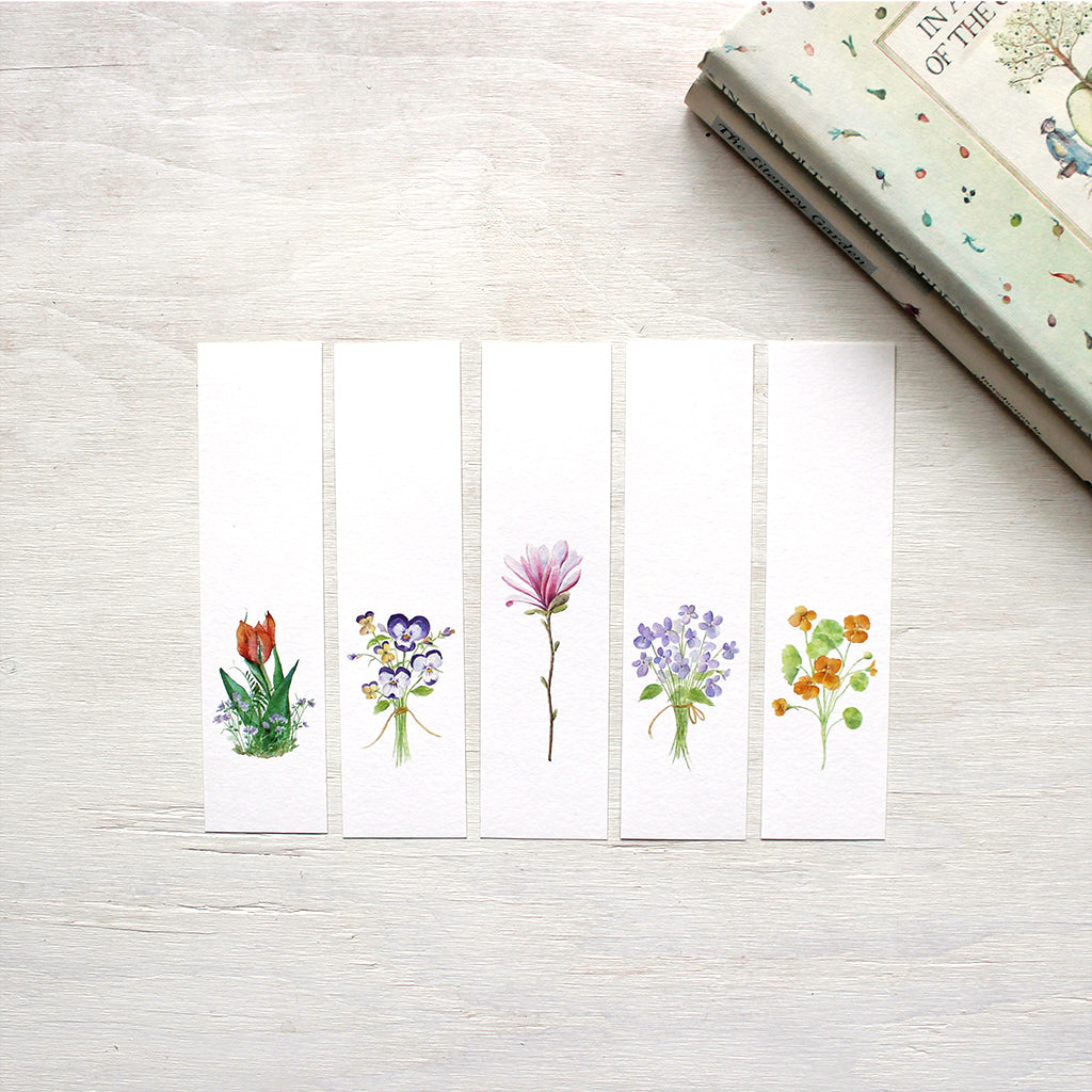 A set of five paper bookmarks featuring watercolor paintings of a tulip and violets, a posy of pansies, a pink star magnolia blossom, a bouquet of violets, and nasturtiums. Artist Kathleen Maunder.