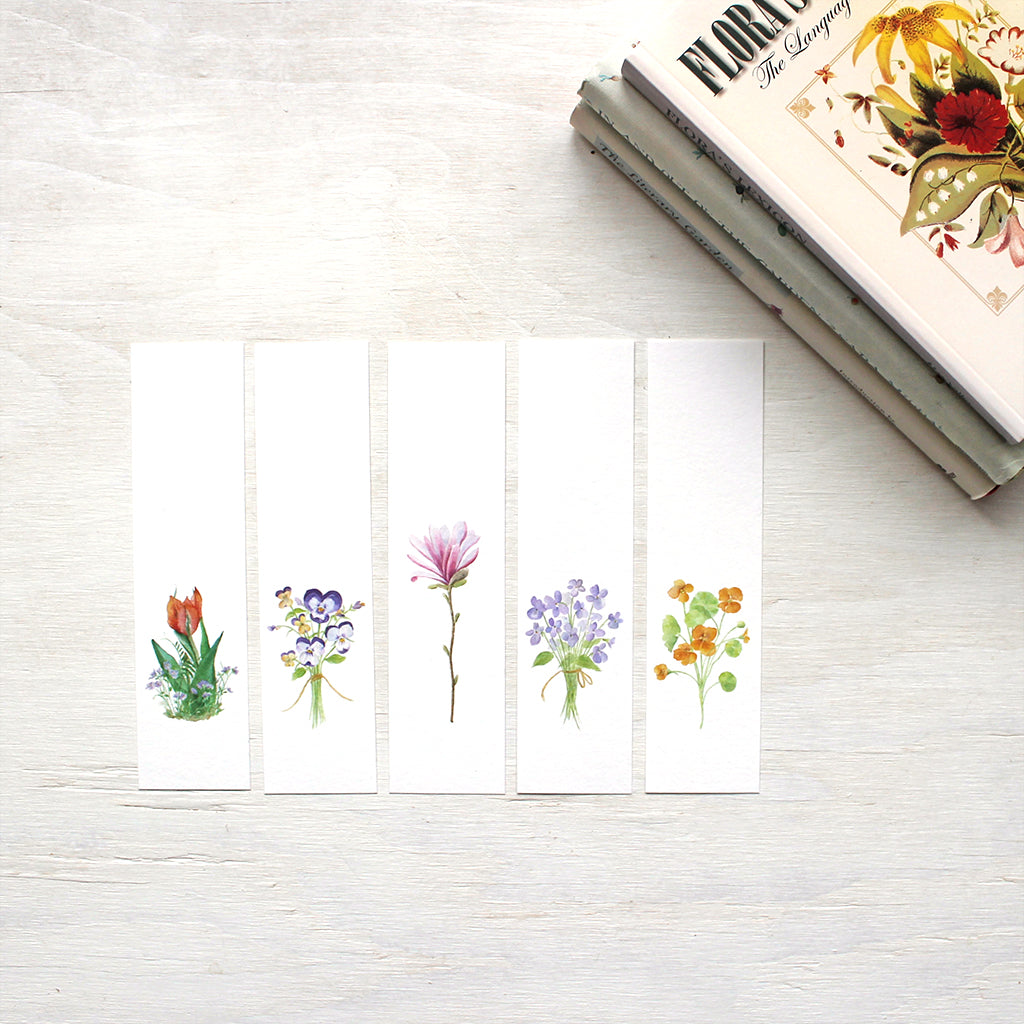 A set of five botanical bookmarks featuring watercolor paintings of a tulip and violets, a posy of pansies, a pink star magnolia blossom, a bouquet of violets, and nasturtiums. Artist Kathleen Maunder.