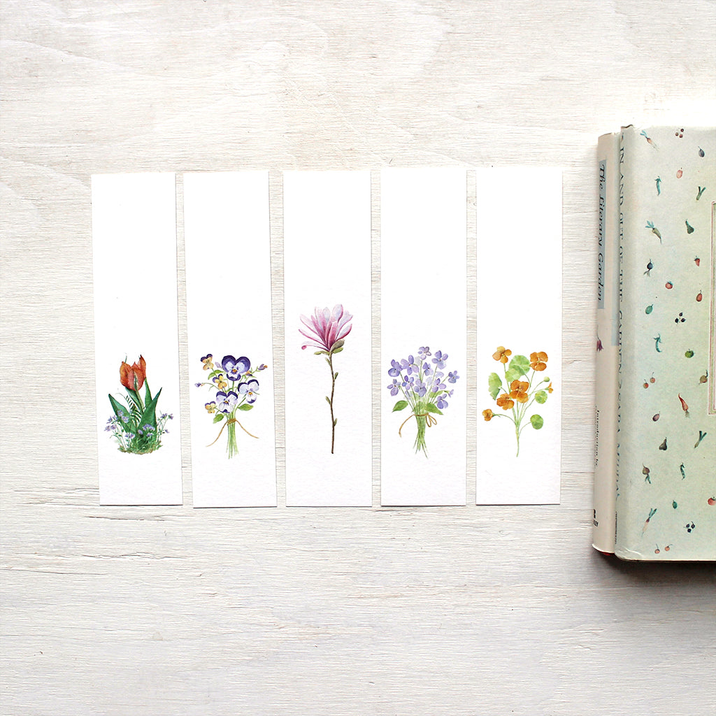 A set of five floral bookmarks featuring watercolor paintings of a tulip and violets, a posy of pansies, a pink star magnolia blossom, a bouquet of violets, and nasturtiums. Artist Kathleen Maunder.