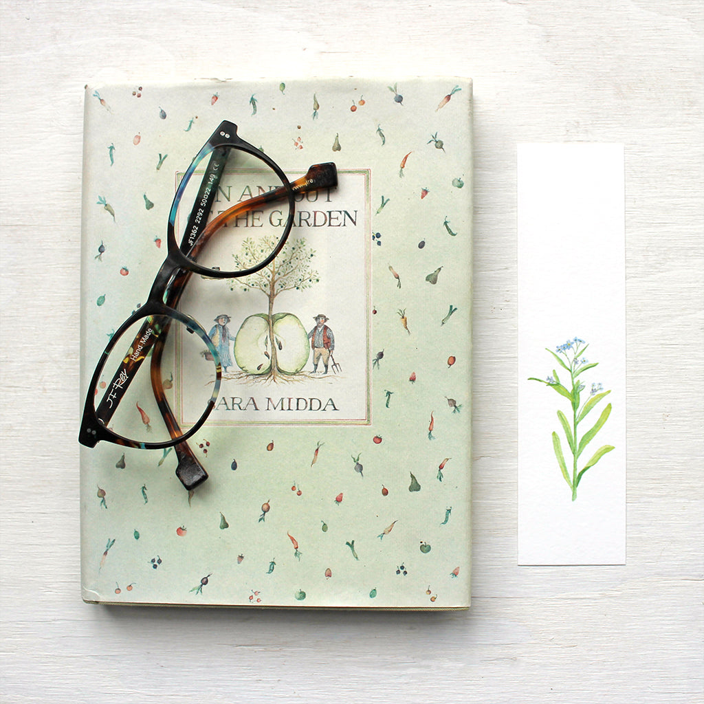 A paper bookmark featuring a watercolor painting of a stem of blue forget me nots. Artist Kathleen Maunder.