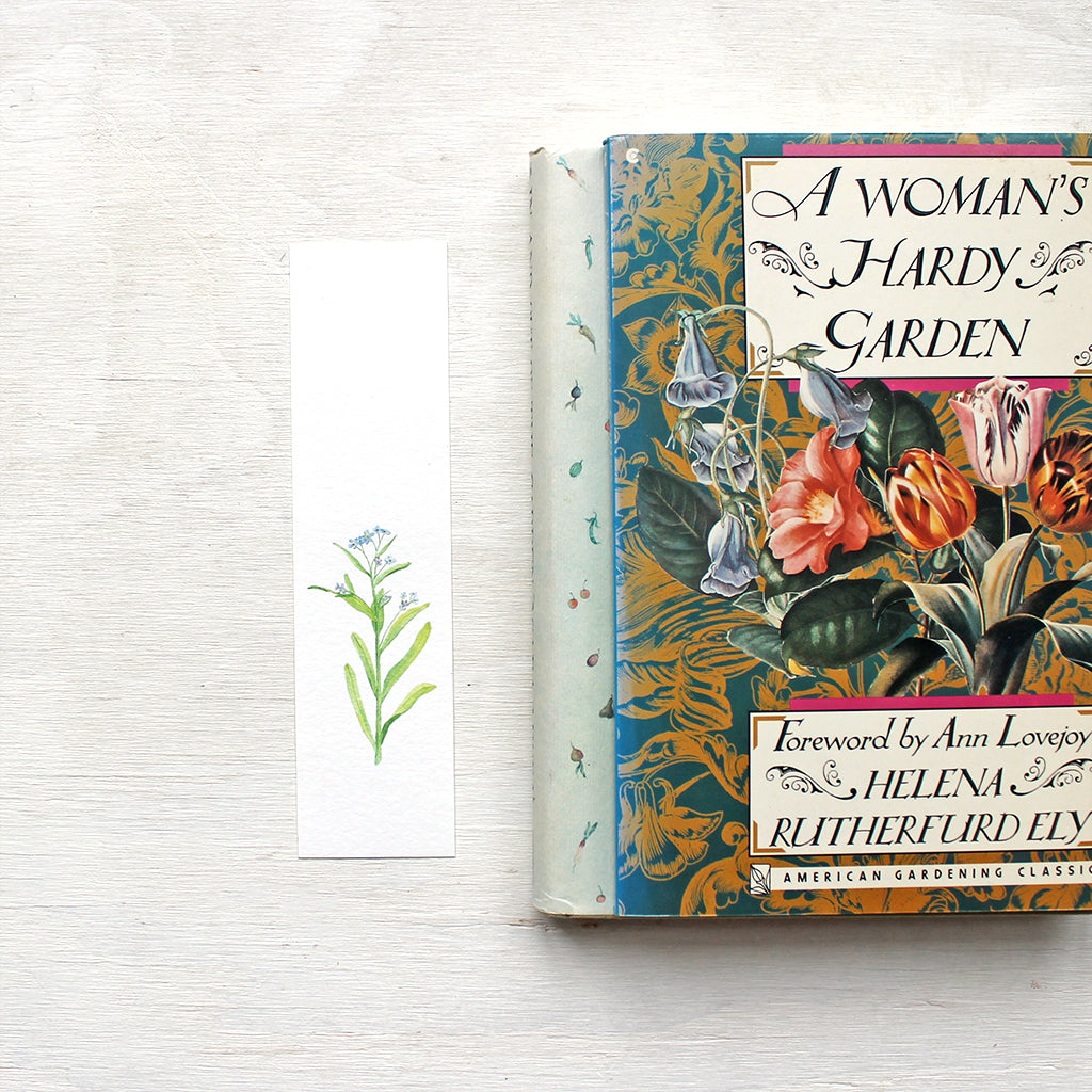 A paper bookmark featuring a watercolour painting of a stem of blue forget me nots. Artist Kathleen Maunder.