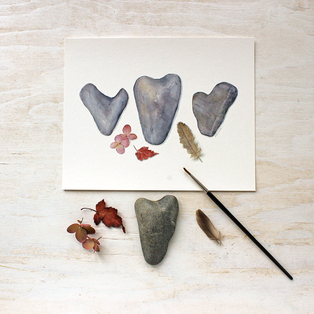 Fragility and Fortitude. A watercolor by Kathleen Maunder depicting three heart shaped rocks, a feather, leaf and hydrangea blossom. Available as an print.