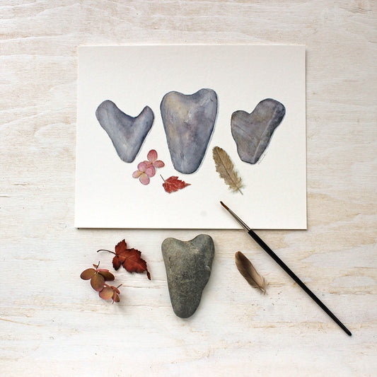 Fragility and Fortitude. A watercolor by Kathleen Maunder depicting three heart shaped rocks, a feather, leaf and hydrangea blossom. Available as an print.