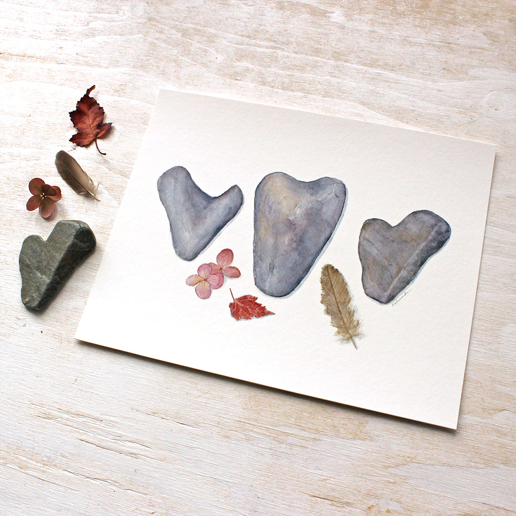 Fragility and Fortitude: A watercolor by Kathleen Maunder depicting three heart shaped rocks, a sparrow feather, leaf and hydrangea blossom. Available as an print.