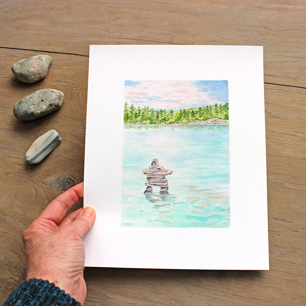 Artist holding a reproduction of a watercolor painting of a lake and inuksuk.