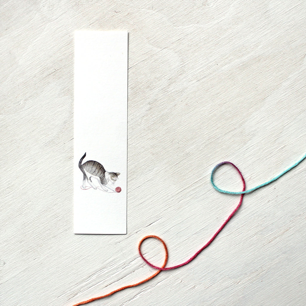 A paper bookmark featuring a watercolor painting of a tabby kitten playing with a ball of yarn. Artist Kathleen Maunder.  