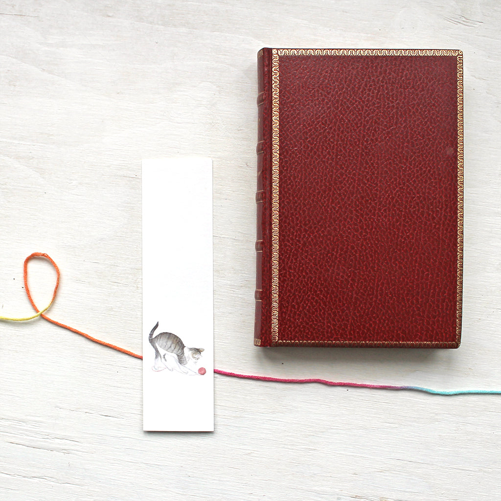 A paper bookmark featuring a watercolor painting of a tabby kitten playing with a ball of yarn. Artist Kathleen Maunder.  (Also shown: an old red leather-covered book.)