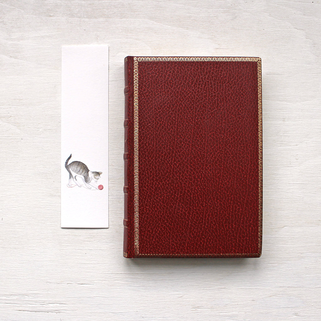 A paper bookmark featuring a watercolor painting of a tabby kitten playing with a ball of yarn. Artist Kathleen Maunder. (Also shown: an old red leather-covered book.)
