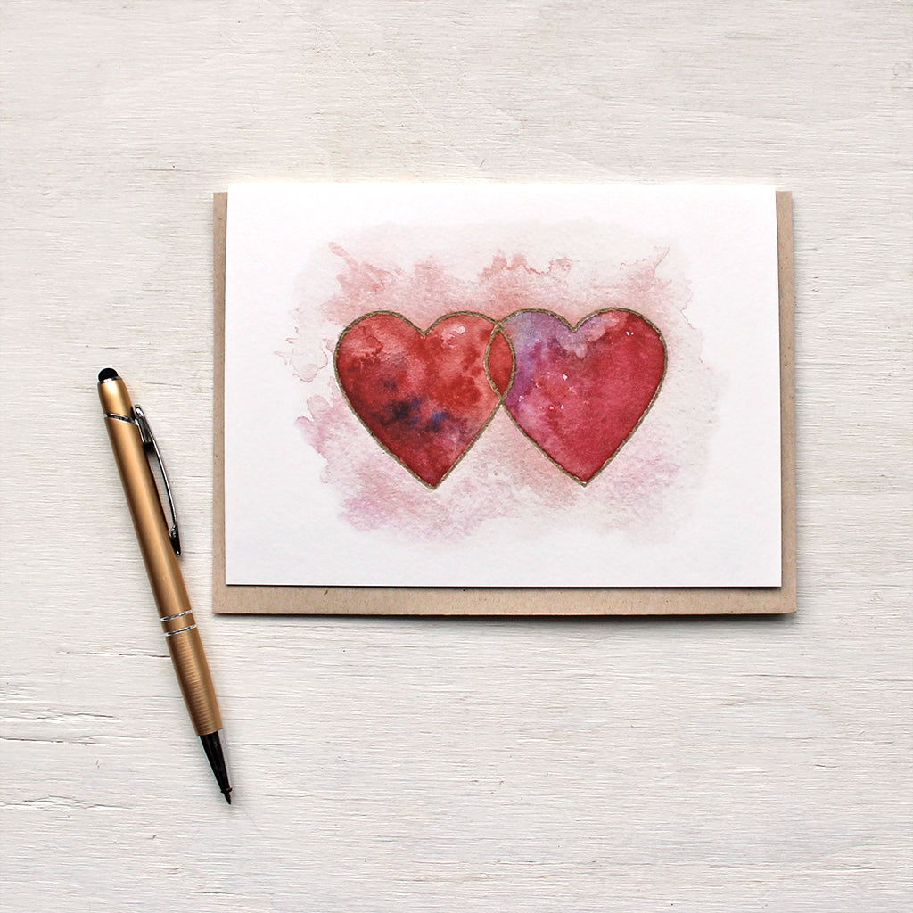 A note card featuring a watercolor painting of two interlocked hearts in tones of red, pink and purple, edged in gold. Artist Kathleen Maunder.