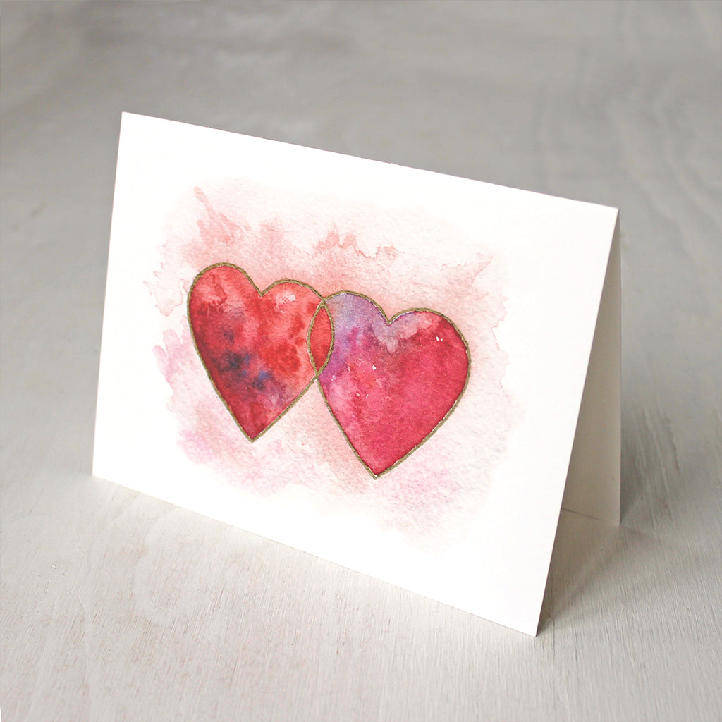 A note card featuring a watercolor image of two interlocked hearts in tones of red, pink and purple, edged in gold. Artist Kathleen Maunder.