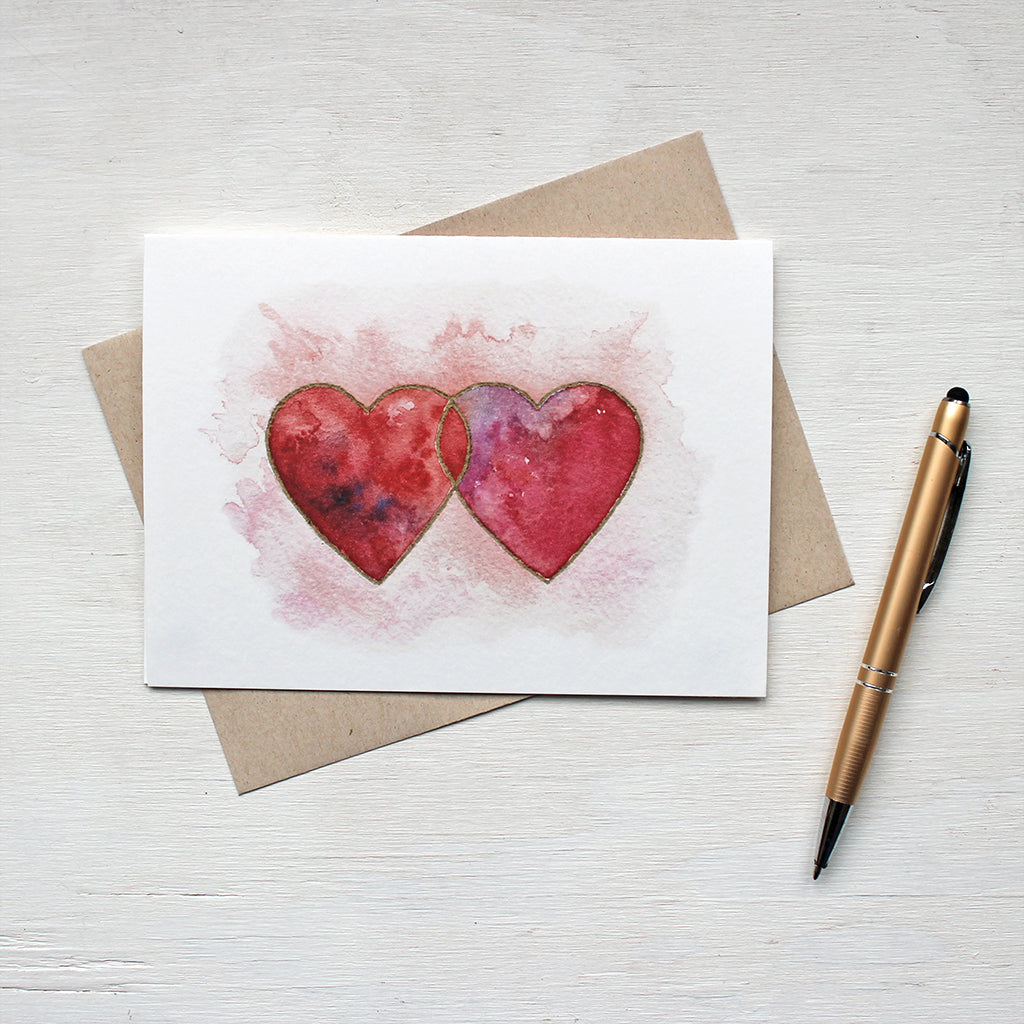 A note card featuring a watercolour image of two interlocked hearts in tones of red, pink and purple, edged in gold. Artist Kathleen Maunder.