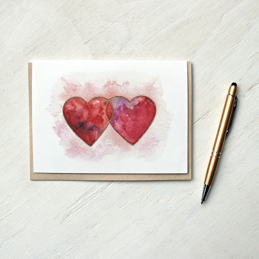 A note card featuring a watercolor image of two interlocked hearts in beautiful tones of red, pink and purple, edged in gold. Artist Kathleen  Maunder.