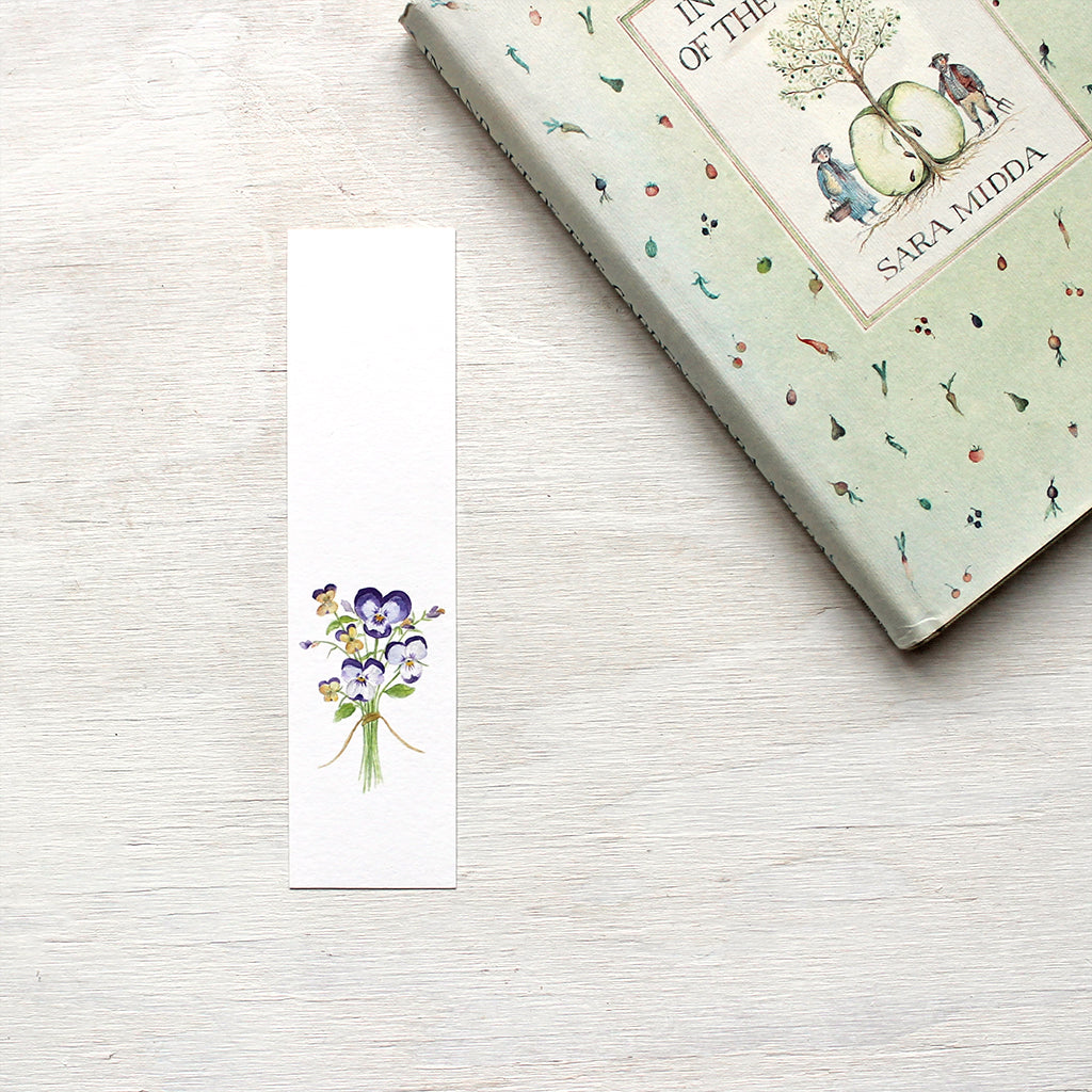 A paper bookmark featuring a watercolor painting of a tiny bouquet of pansies and violas. Artist Kathleen Maunder.