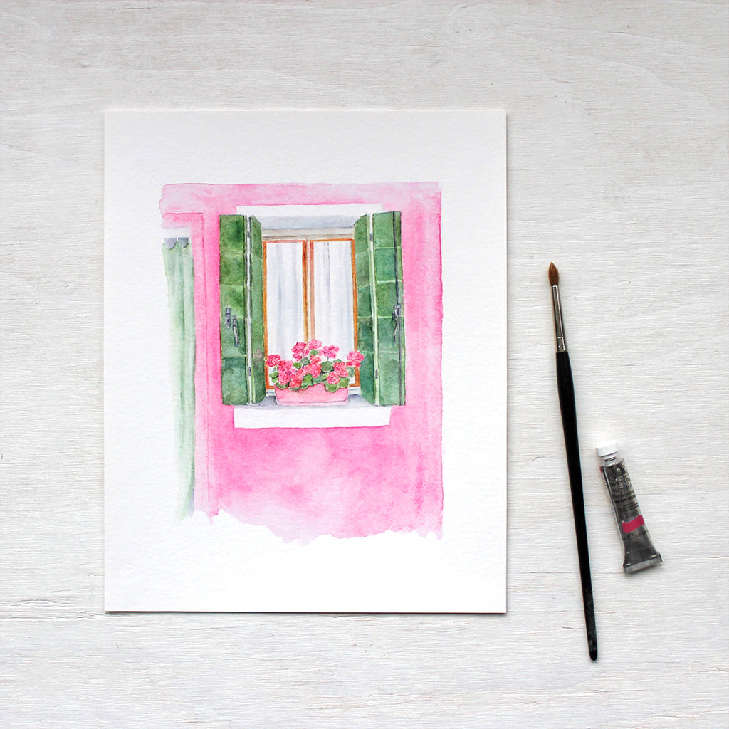 An watercolor art print depicting a pink house with green shutters and a pot of geraniums. A scene from Burano, Italy. Artist Kathleen Maunder